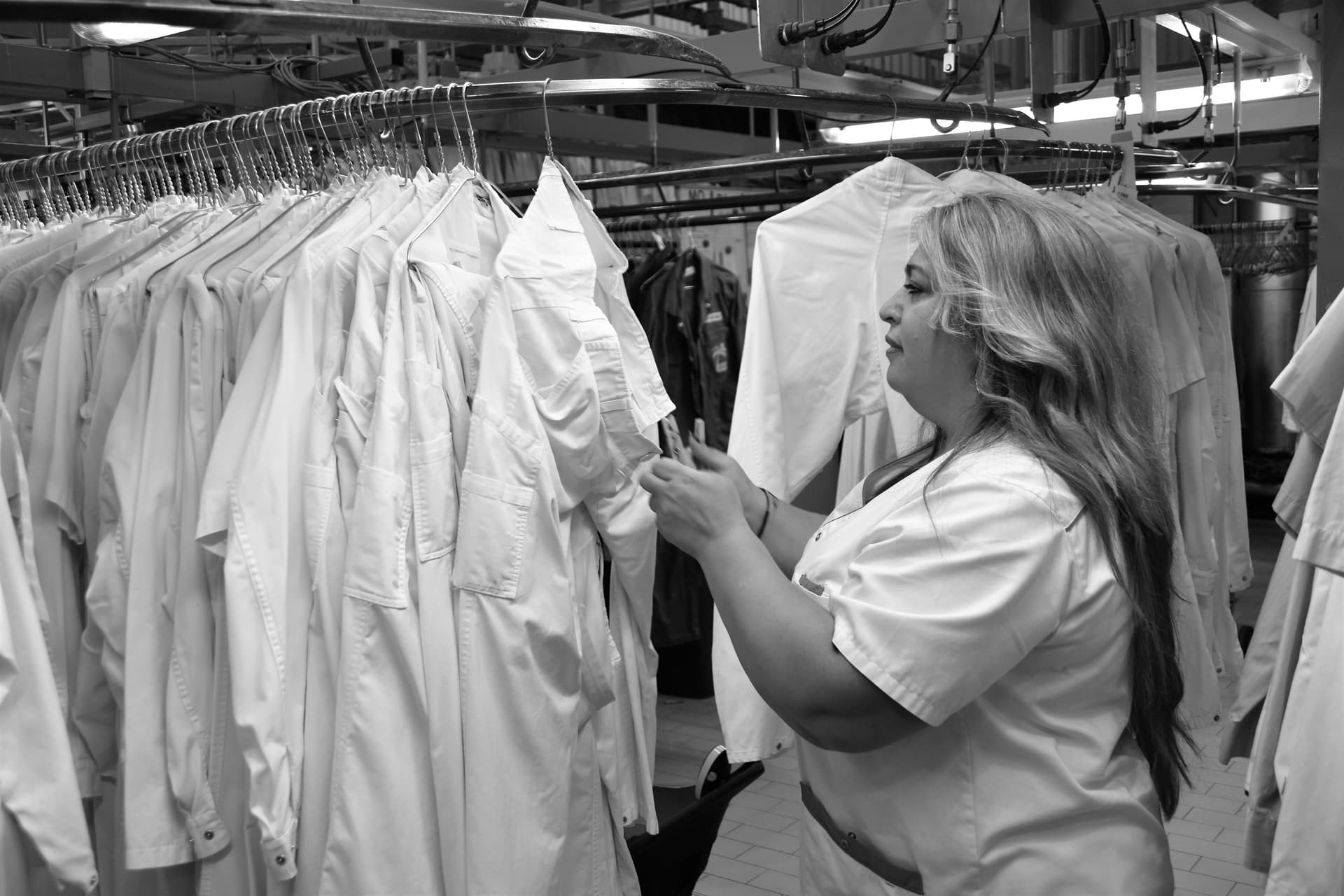 Professional laundry worker in white uniform carefully inspecting blue dress shirt under bright lighting